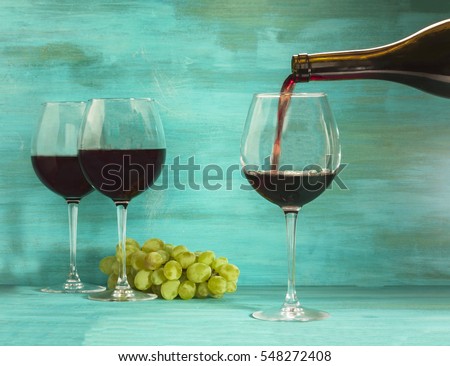 Photo of red wine being poured into glass from bottle, with more glasses and grapes in the background, on vibrant turquoise with copyspace