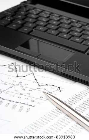 Business analytics workplace - sheet with numbers, graph, pen, and laptop, selective focus on pen