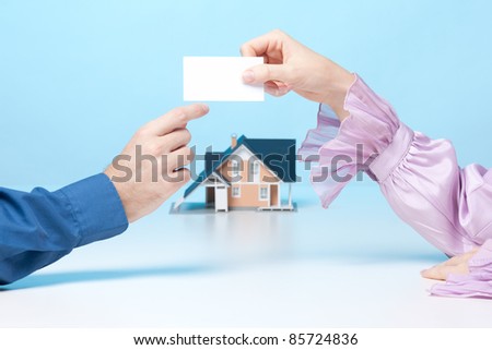 Real estate broker woman is giving to client her business card. Real estate agency is represented by model of house in background.