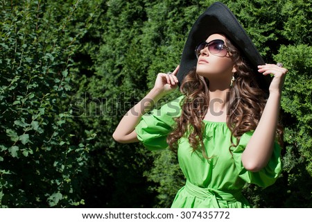 Model with long curly hair in a park holding hands hat. The girl in the green dress, black hat and sunglasses