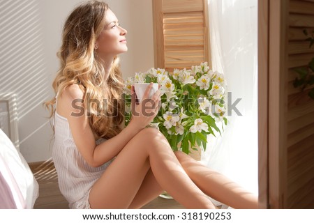 The sun is shining into the bedroom, she looks out the window. The lady sitting beside the bed, her hand near her head, she looks into the camera. Morning coffee, enjoyment, she closed her eyes