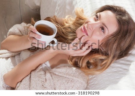 The sun is shining into the bedroom, she looks out the window. The lady sitting beside the bed, her hand near her head, she looks into the camera. Morning coffee, enjoyment, she closed her eyes