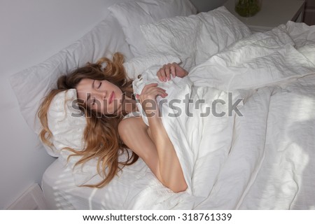 Women\'s head with curly hair, light make-up on her face. The sun shines in the bedroom and in the face of a woman. Beautiful woman with closed eyes in her bed woman sleeping on the back. Woman sleeps
