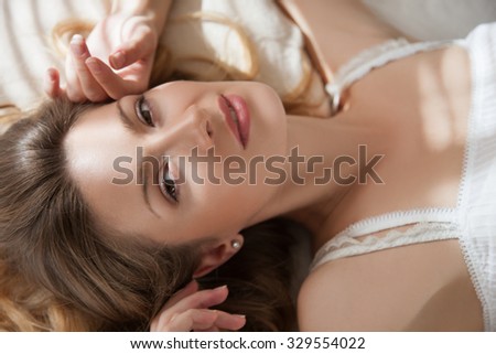 Female portrait close up lying on the bed in the bedroom the sun shines through the window.seductive woman.woman and bedroom.woman close-up looks at  camera brown eyes lying on her back.morning in bed