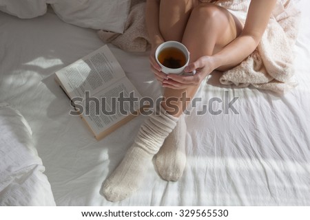 Long female feet in warm woolen socks around her feet is a book she is reading it.Part of the body.Beautiful female legs sitting on the bed with a cup in his hands.Morning with a book in their hands.