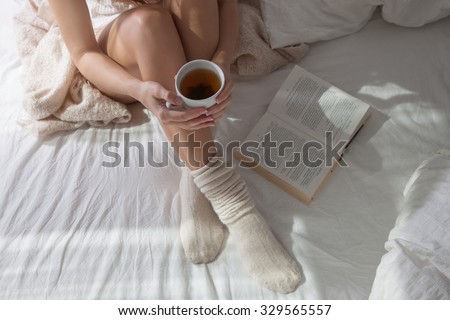 Shapely legs sitting on a bed with a light bed in a warm and cozy bed. A woman sitting on a bed in her hands a cup of hot tea, she reads a book. long female legs in socks