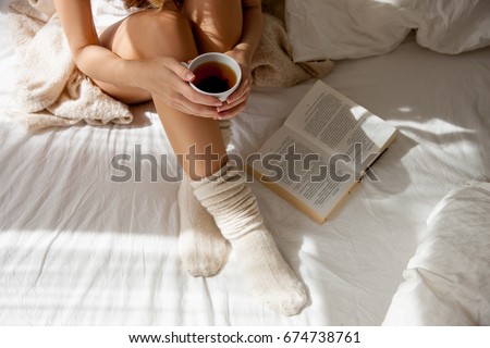 Beautiful Legs close-up in Bed. Woman is Drinking Tea and Reading a Book. Girl sitting on a Bed in Woolen socks. Beautiful Woman no Face and Legs. Attractive Model Wears woolen white socks
