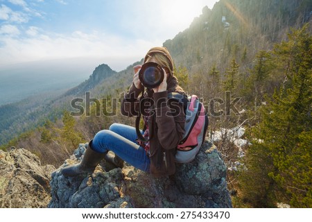 Adventurous female photographer sitting on the rock while photographing mountains facing the off scene viewer against the setting sun. Wide angle perspective. Tourism, adventure, hiking concept.