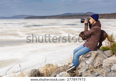 Female photographer sitting one the boulder near her dog against mountain landscape at a frozen lake while shooting