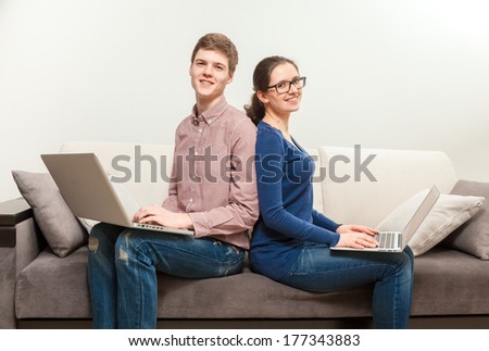 Portrait of young woman and man sitting back to back on couch and holding laptops