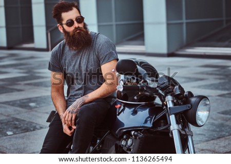 Brutal bearded male in sunglasses dressed in a gray t-shirt and black pants sitting on his custom-made retro motorcycle against a skyscraper.