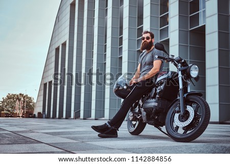 Brutal bearded male in a gray t-shirt and black pants holds a helmet sitting on his custom-made retro motorcycle against skyscraper.