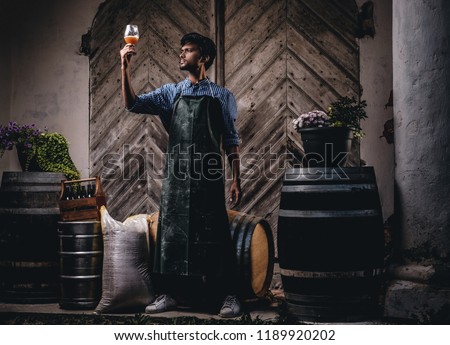 Expert brewer in apron holds glass of beer and checking quality of brewed drink at brewery factory.