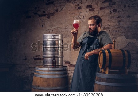 Bearded brewer in apron standing near barrels checking the quality of brewed drink at old brewery factory.
