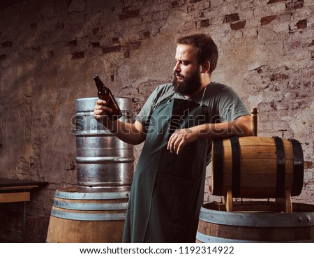 Brewer in apron standing near barrels and drinks craft beer at old brewery factory.