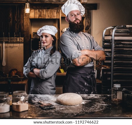 Chef with his assistant in cook uniform posing with crossed arms near table with ready dough in the bakery.