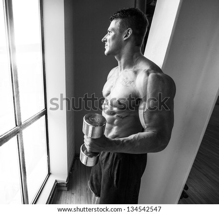 Black and white image of bodybuilder who is staring in window while having a training with dumbbells