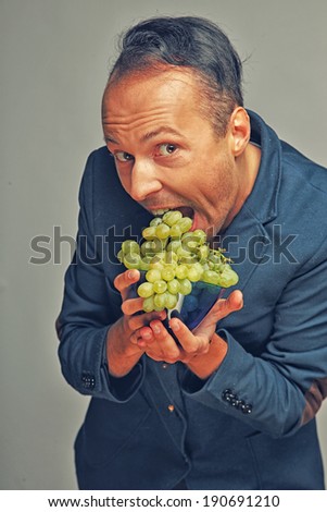 A man wearing a jacket trying to eat a lot of grapes