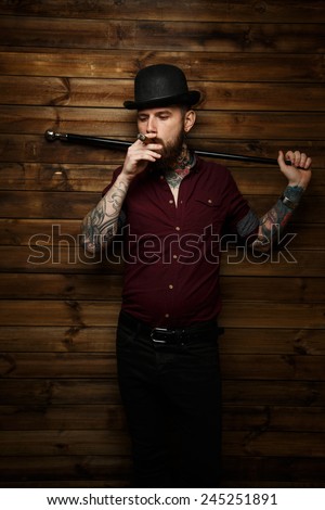 Brutal man with tattooes and hat on his had smoking holding walking stick