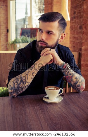 Fashionable male with tattooes sitting at the table in coffee shop with a cup of coffee