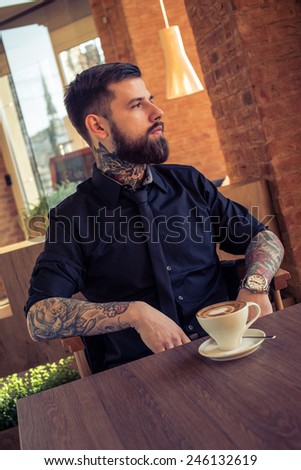 Fashionable male with tattooes sitting at the table in coffee shop with a cup of coffee