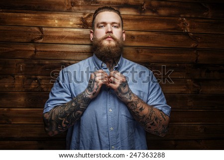 Brutal male with beard and tatooes buttons up his t-shirt Brutal male with beard and tatooes buttons up his t-shirt