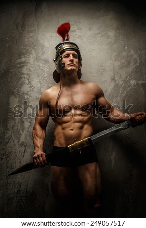 Muscular warrior with sword and helmet posing in front of concrete wall