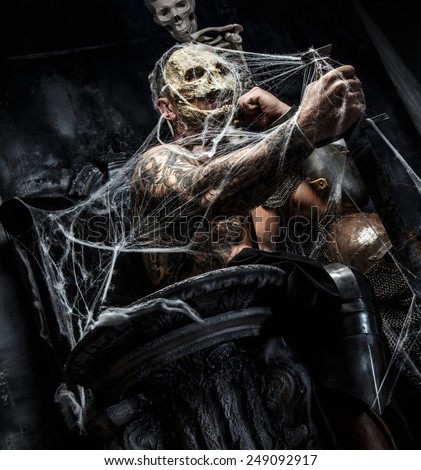 Muscular man with skull on his face and sword in his hand posing on the throne in spiders web on gray background.