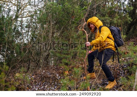 Smiling man with beard in a forest. Dressed in yellow jacket, black bants, yellow boots, with stick and backpack.