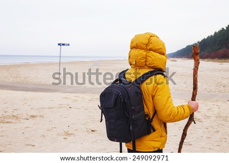 Man from behind dressed in yellow jacket with black backpack, hold stich, stands on the winter beach.