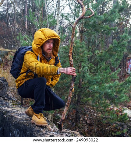 Man with beard  in yellow jacket, yellow boots and black pants with bacpack and stick  walking in a forest.