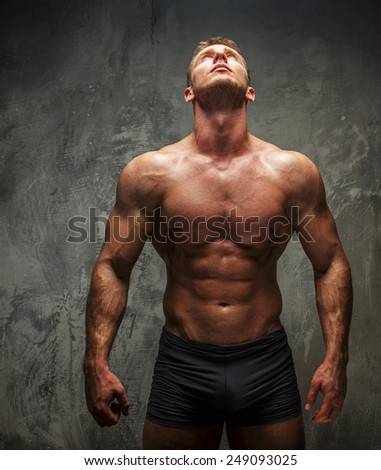 Muscular man with great body relief watching up on white light. Grey background.