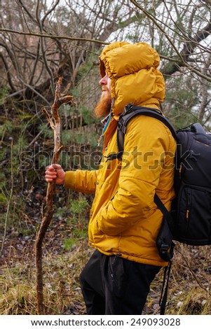 Profile of a man with beard in yellow jacket, black pants and black backpack. In the forest. Hold stick.