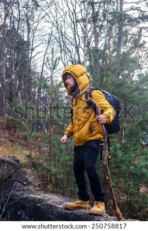 Man with beard  in yellow jacket, yellow boots and black pants with backpack and stick  walking in a forest.