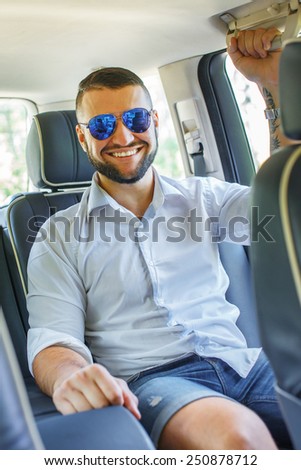 Happy man with black hair and beard, sunglasses, dressed in white shirt and blue shorts sitting on back seat of a car.