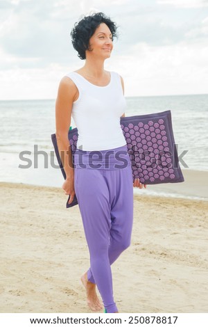 Middle age woman with shirt black hair in white t-shirt and purple pants posing on the summer beach. Rug for massage.