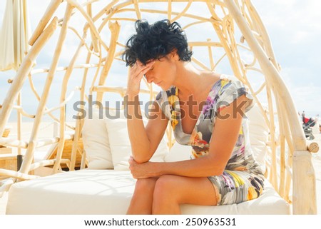 Middle age woman with shirt black hair in color dress sitting on beach sofa with headache.