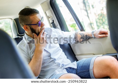 Happy man with black hair and beard, sunglasses, dressed in white shirt and blue shorts sitting on back seat of a car and looking in the car window.