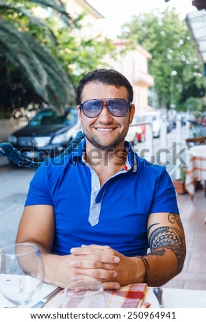 Smiling male in blue t shirt, sun glasses, tattoo on his hand sitting at the cafe table with exotic background.