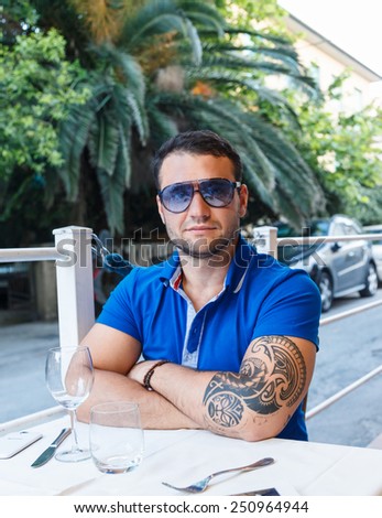 Portrait of a man with tattoo on his hand. Wearing sun glasses, dressed in blue t shirt, sitting at a cafe table in exotic country.