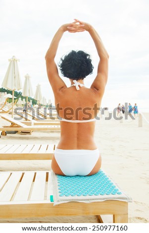 Middle age woman with black hair in white bikini sitting on a beach sofa and doing yoga. Azure rug for massage.