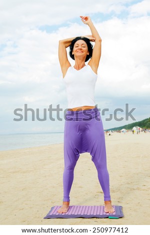 Middle age woman with shirt black hair in a white t-shirt and purple pants doing yoga on the summer beach.