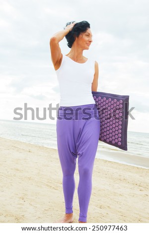 Middle age woman with shirt black hair in white t-shirt and purple pants posing on the summer beach. Rug for massage.
