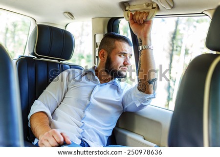 Happy man with black hair and beard, sunglasses, dressed in white shirt and blue shorts sitting on back seat of a car and looking in the car window.