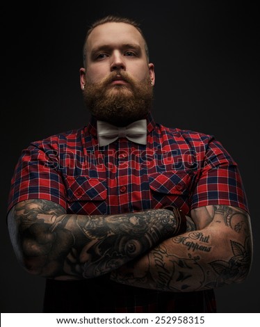 Portraite of brutal hipster with tattooes on his hand dressed in red shirt and bow tie.