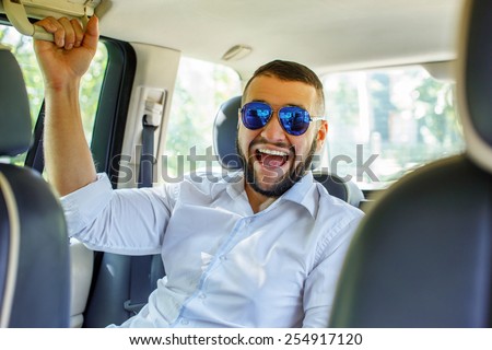 Happy man with black hair and beard, sunglasses, dressed in white shirt and blue shorts sitting on back seat of a car.