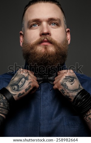 Male with beard and tatoos in blue shirt and bow tie. Isolated on dark grey background.