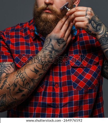 Huge brutal man with tattoo shawing his beard with a razor