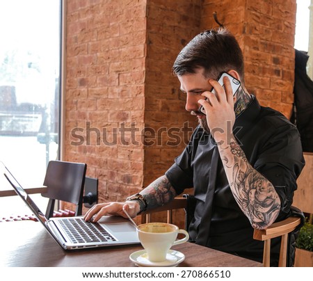 Handsome modern man calling by smartphone and working with laptop. Cafe interior.