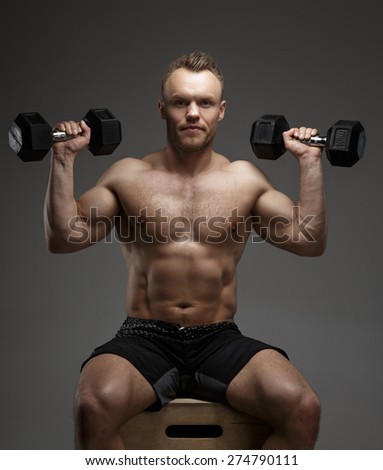 Muscular bodybuilder guy sitting on chair and doing exercises with dumbell on grey background